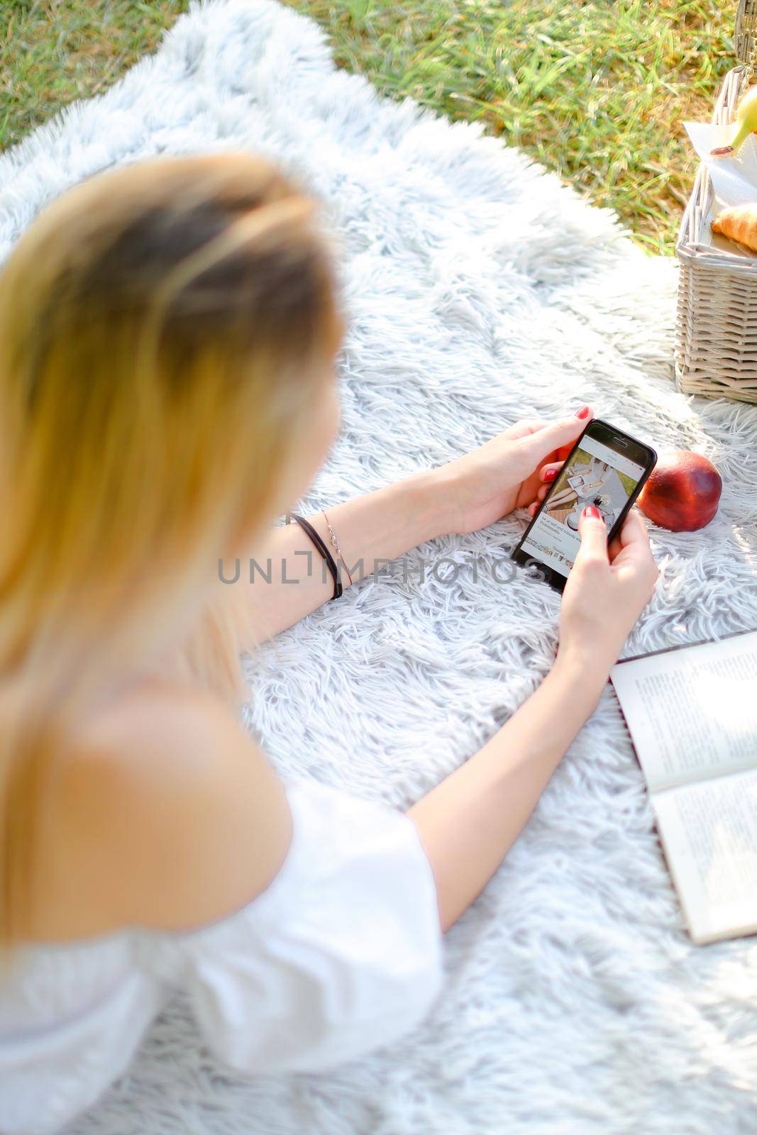 Young caucasian girl with red nails lying in park on plaid and enjoying social networks by smartphone. Concept of having free time, picnic and modern technology.