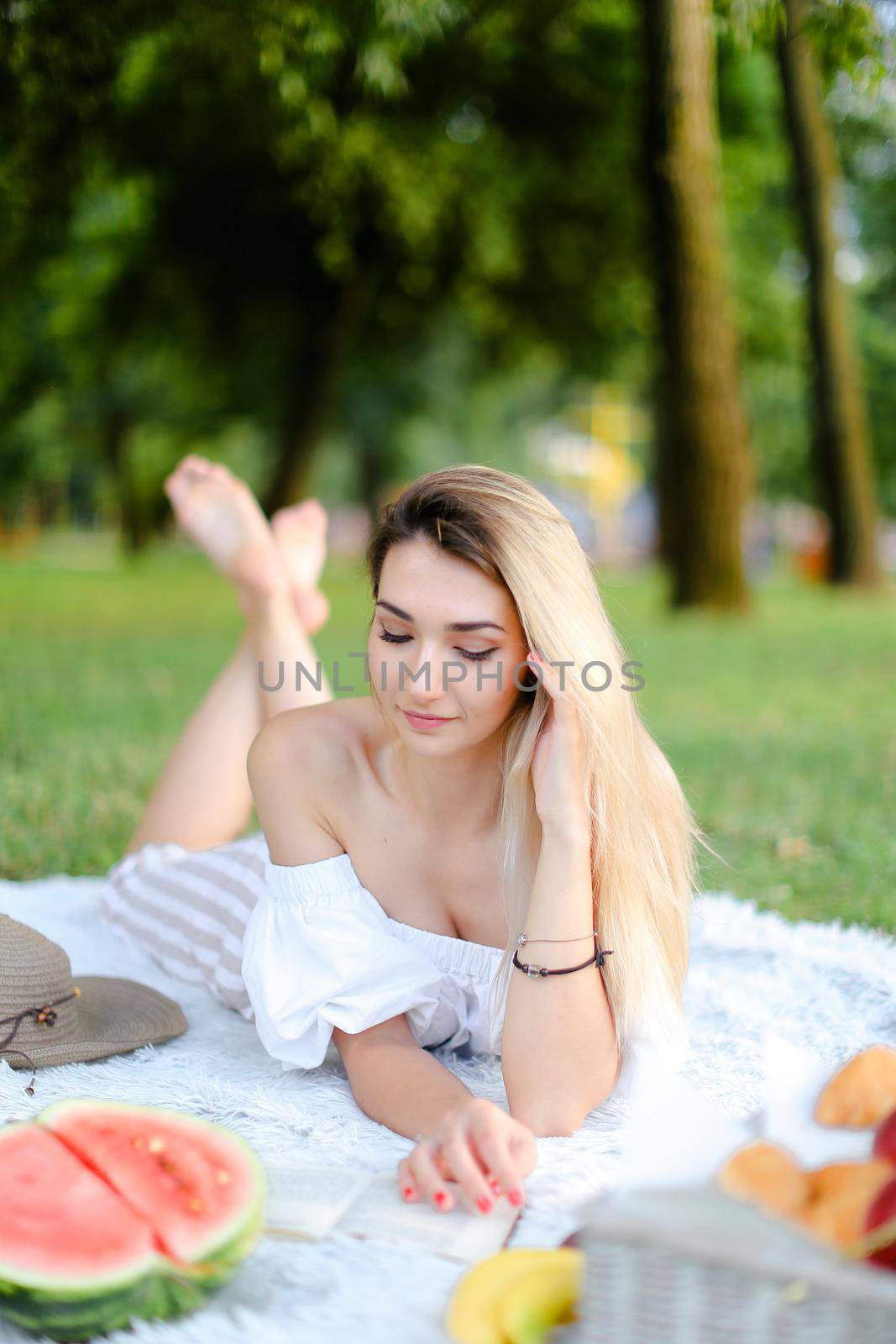 Young caucasian girl lying on plaid near watermelon in park and reading book. Concept of picnic on nature, summer vacations and leisure time.