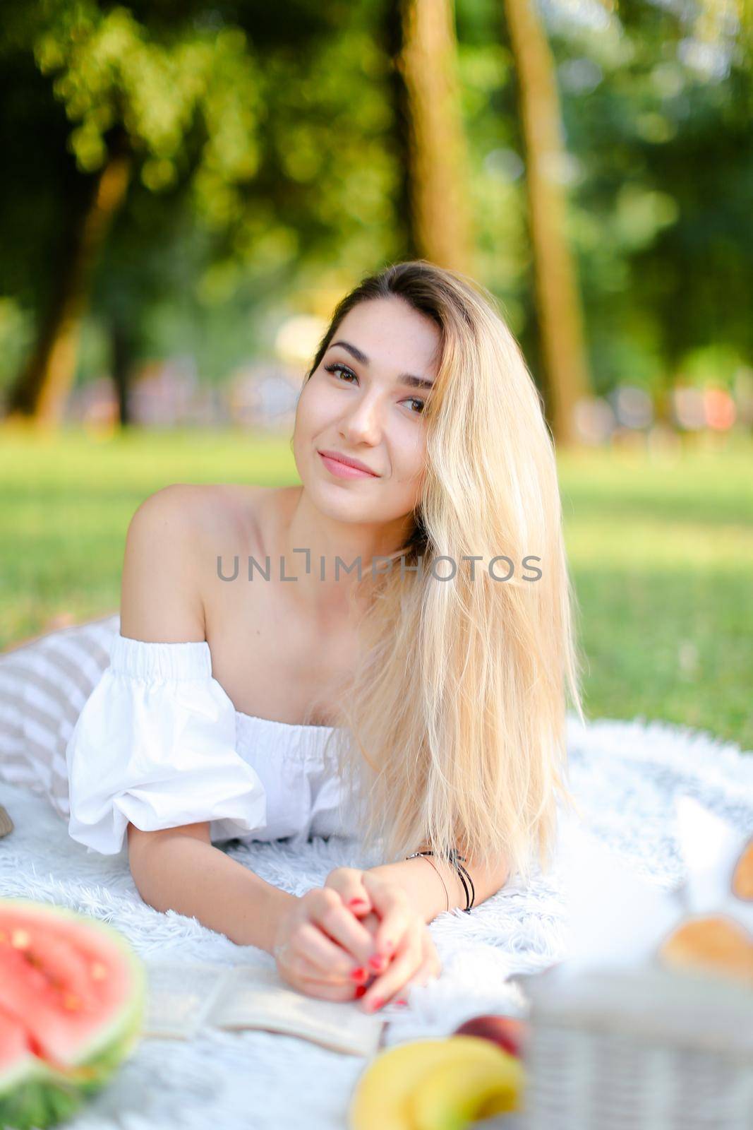 Young european woman lying on plaid near watermelon in park and reading book. Concept of picnic on nature, summer vacations and leisure time.