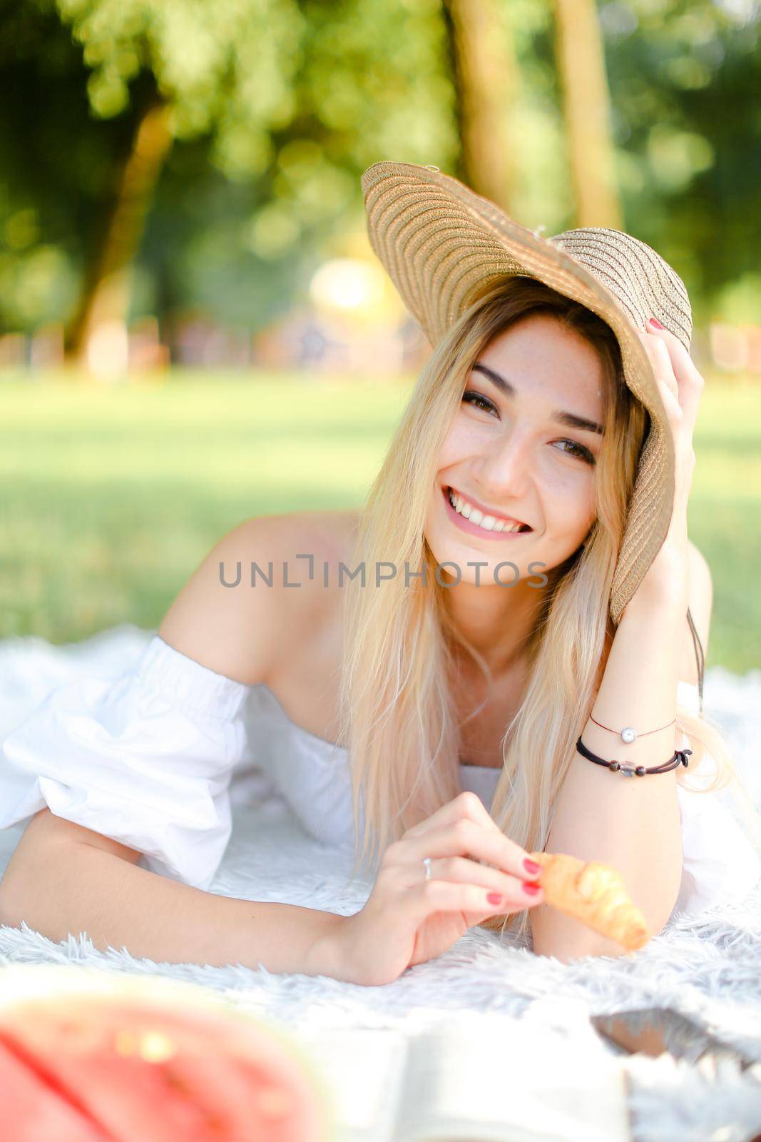 Young caucasian woman in hat keeping croissant, lying in park on plaid and reading novel. Concept of picnic and interesting book, bakery products.