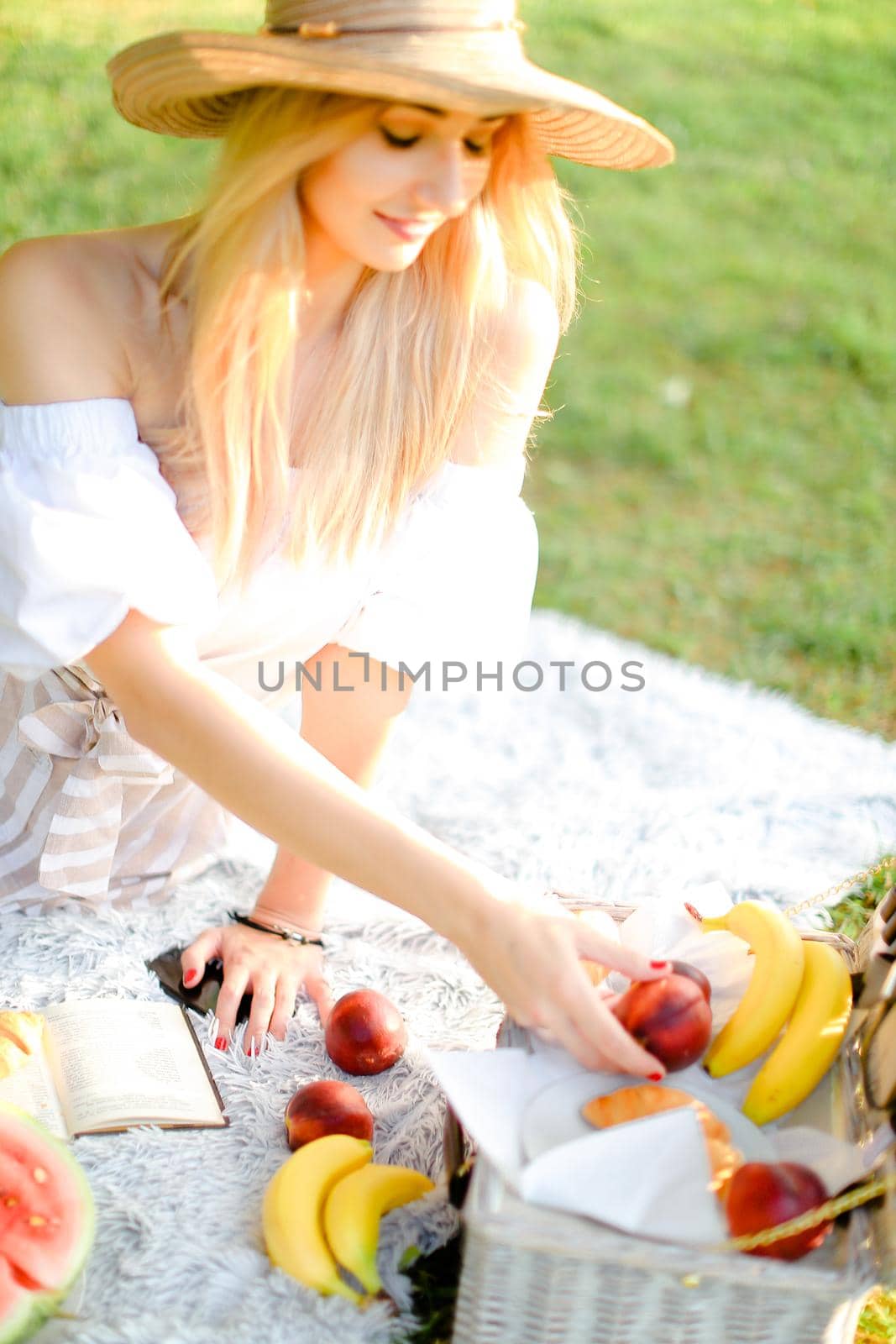 Young girl in hat eating fruits and lying on plaid with book, grass in backrground. Concept of healthy food, picnic and summer season.