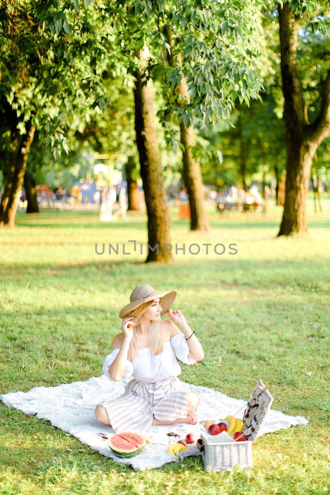 Young european woman in hat sitting in park on plaid near fruits, grass and trees in background. Concept of summer picnic, resting on nature and healthy food.