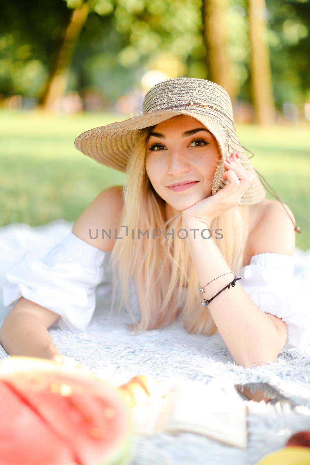 Young caucasian woman in hat lying in park on plaid and reading novel. Concept of picnic and interesting book, resting in park on open air.