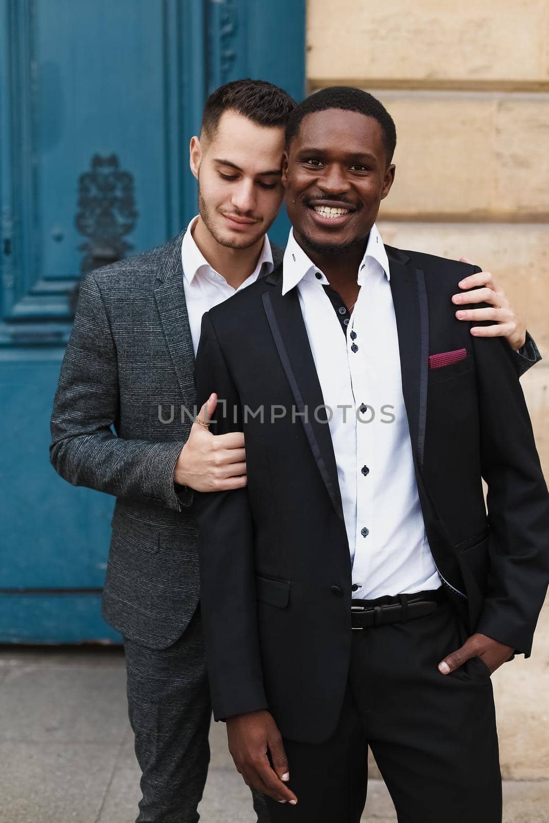 Two boys, caucasian and afro american, wearing suits standing near building and hugging. Concept of gays and lgbt.