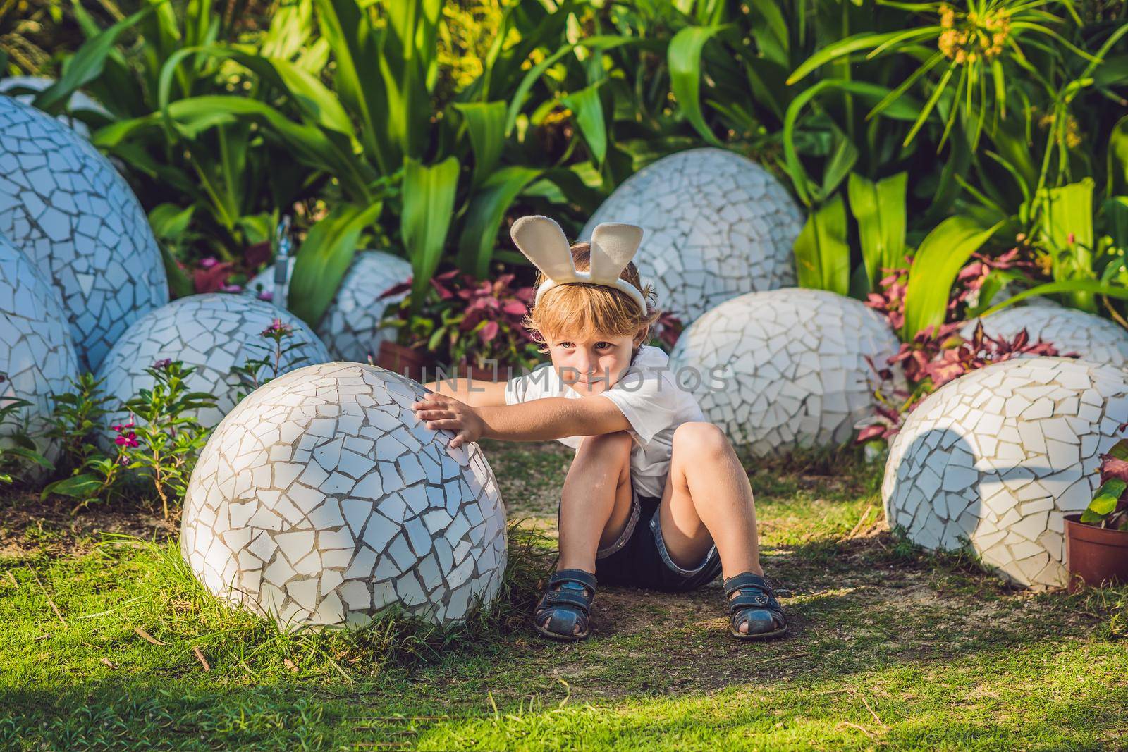 Cute little kid boy with bunny ears having fun with traditional Easter eggs hunt, outdoors. Celebrating Easter holiday. Toddler finding, colorful eggs by galitskaya