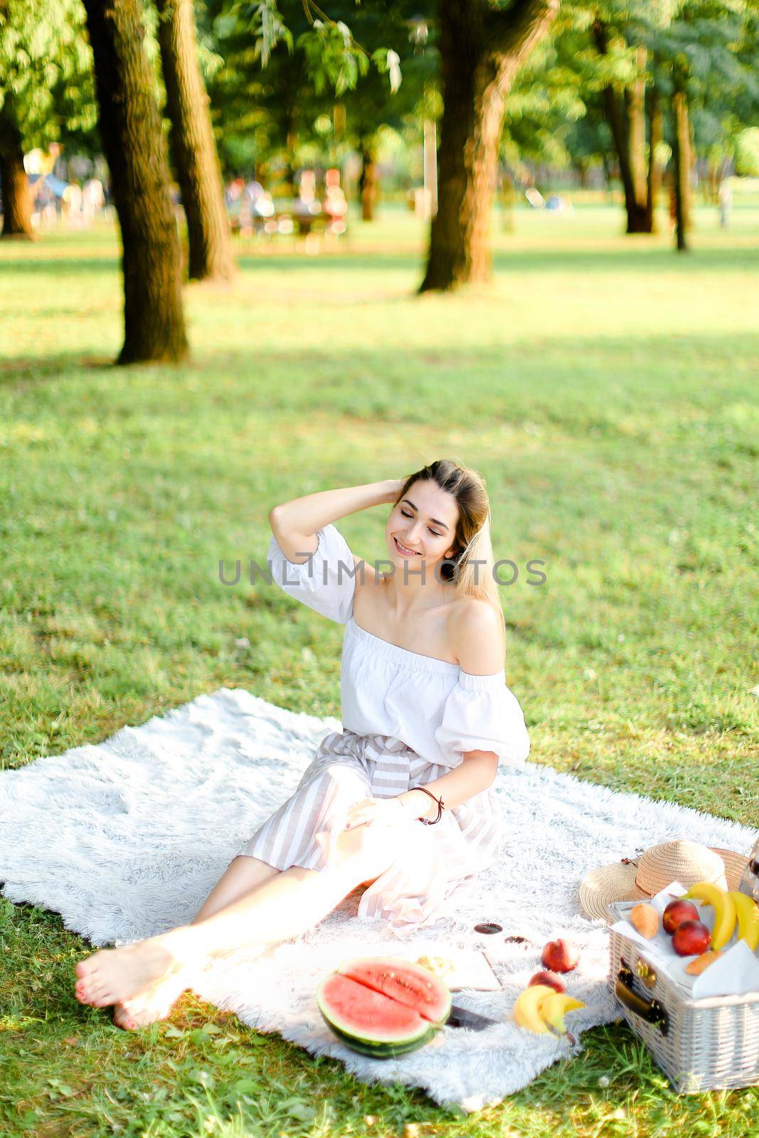 Young girl sitting on plaid with fruits in park. Concept of summer picnic, photo session in open air and vacations.