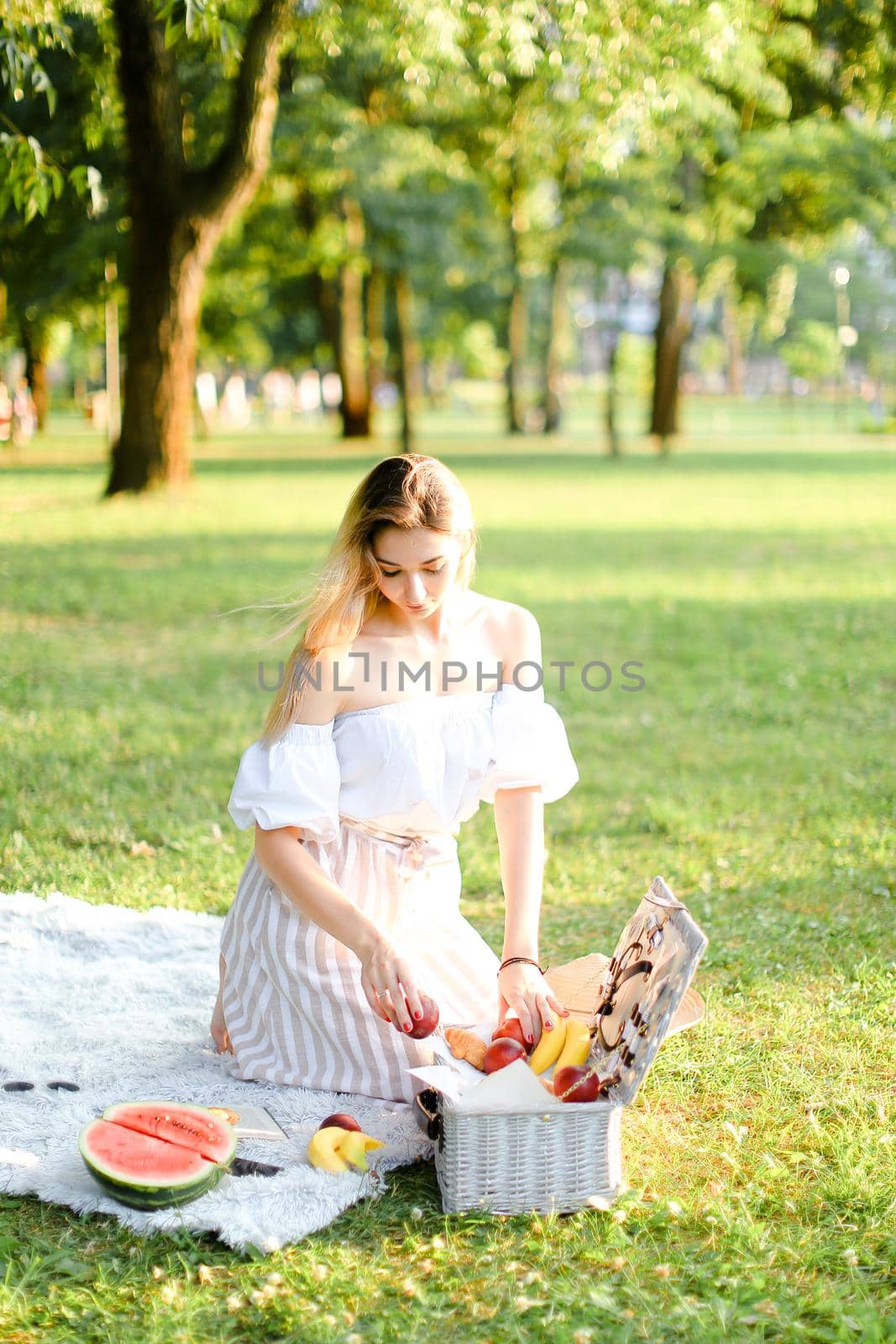 Young blonde woman preparing for picnic in park, sitting on plaid near box and waterlemon. Concept of having leisure time, resting in open air and summer season.