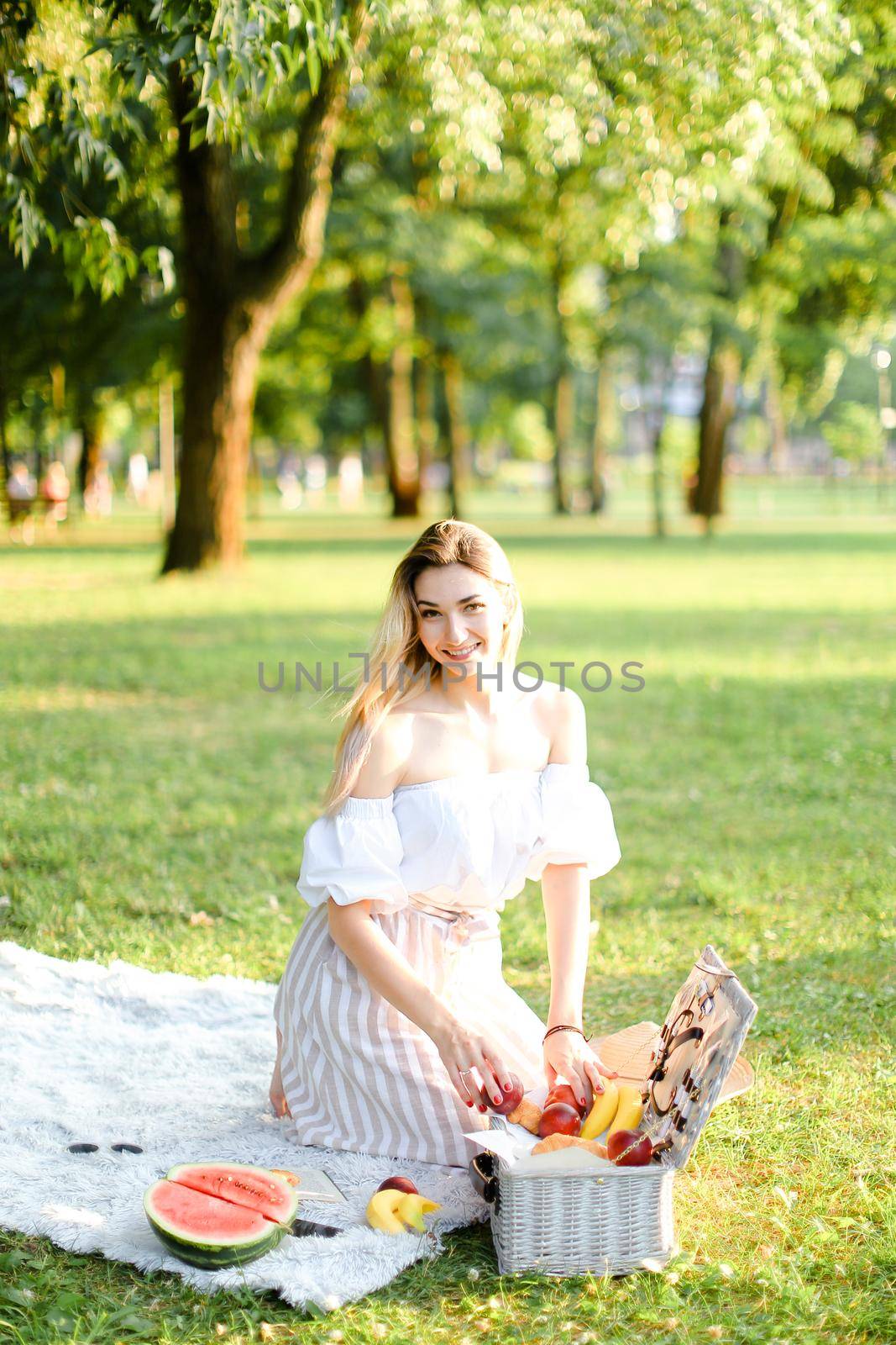 Young happy woman preparing for picnic in park, sitting on plaid near box and waterlemon. Concept of having leisure time, resting in open air and summer season.