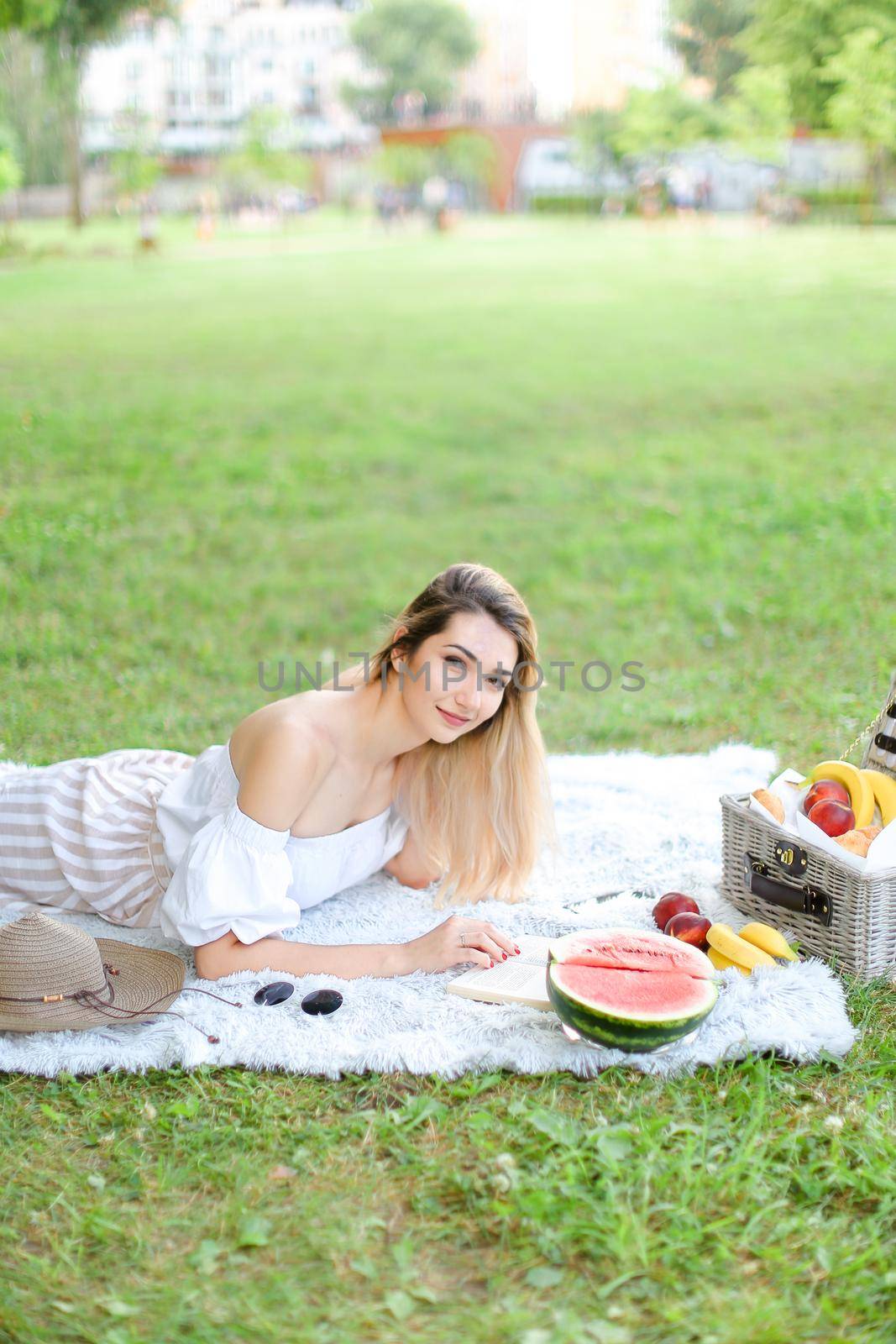 Young pretty blonde woman reading book and lying on plaid near fruits and hat, grass in background. Concept of summer picnic and resting on weekends in open air.