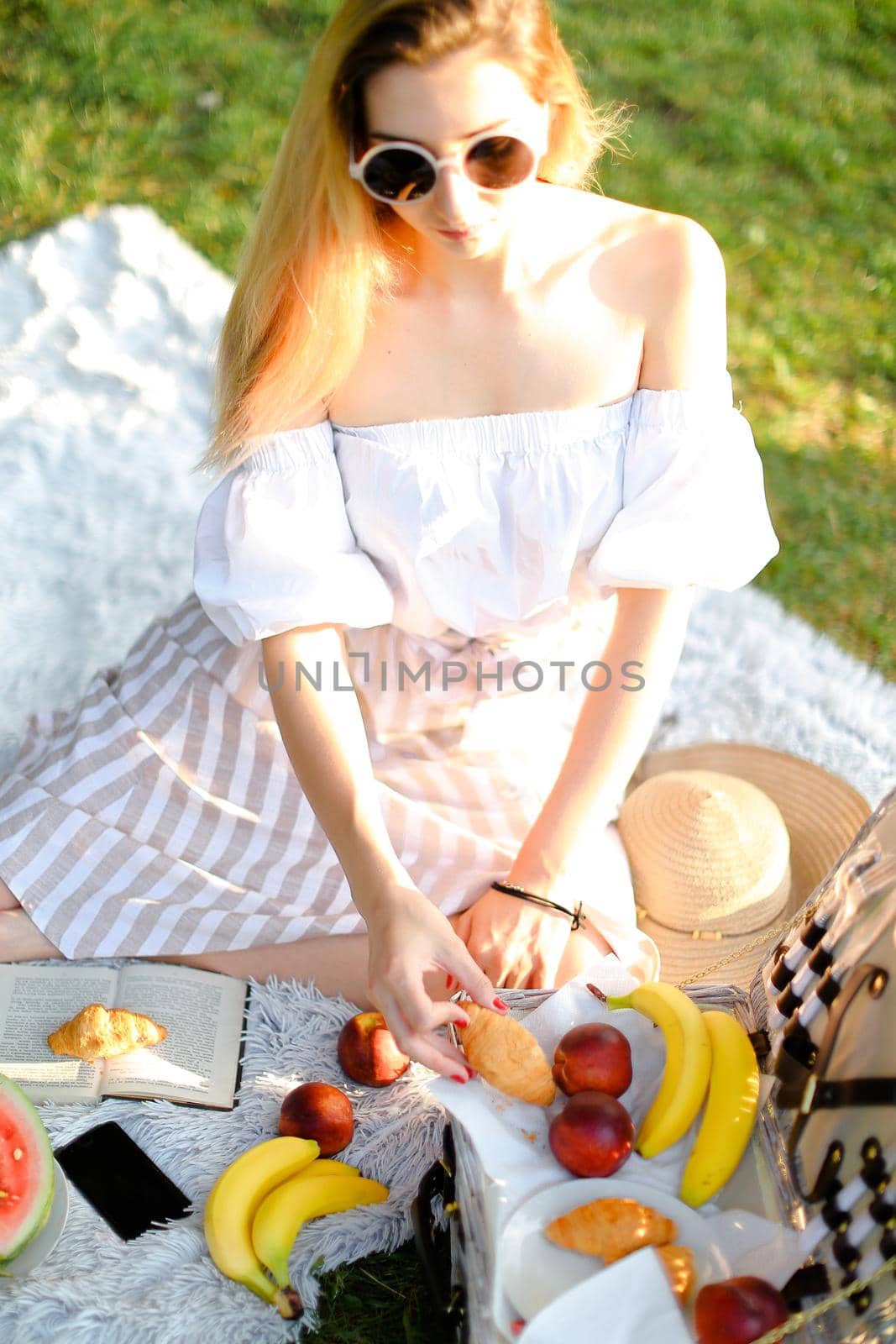 Young girl in sunglasses having picninc and sitting on plaid near book, fruits and smartphone. by sisterspro