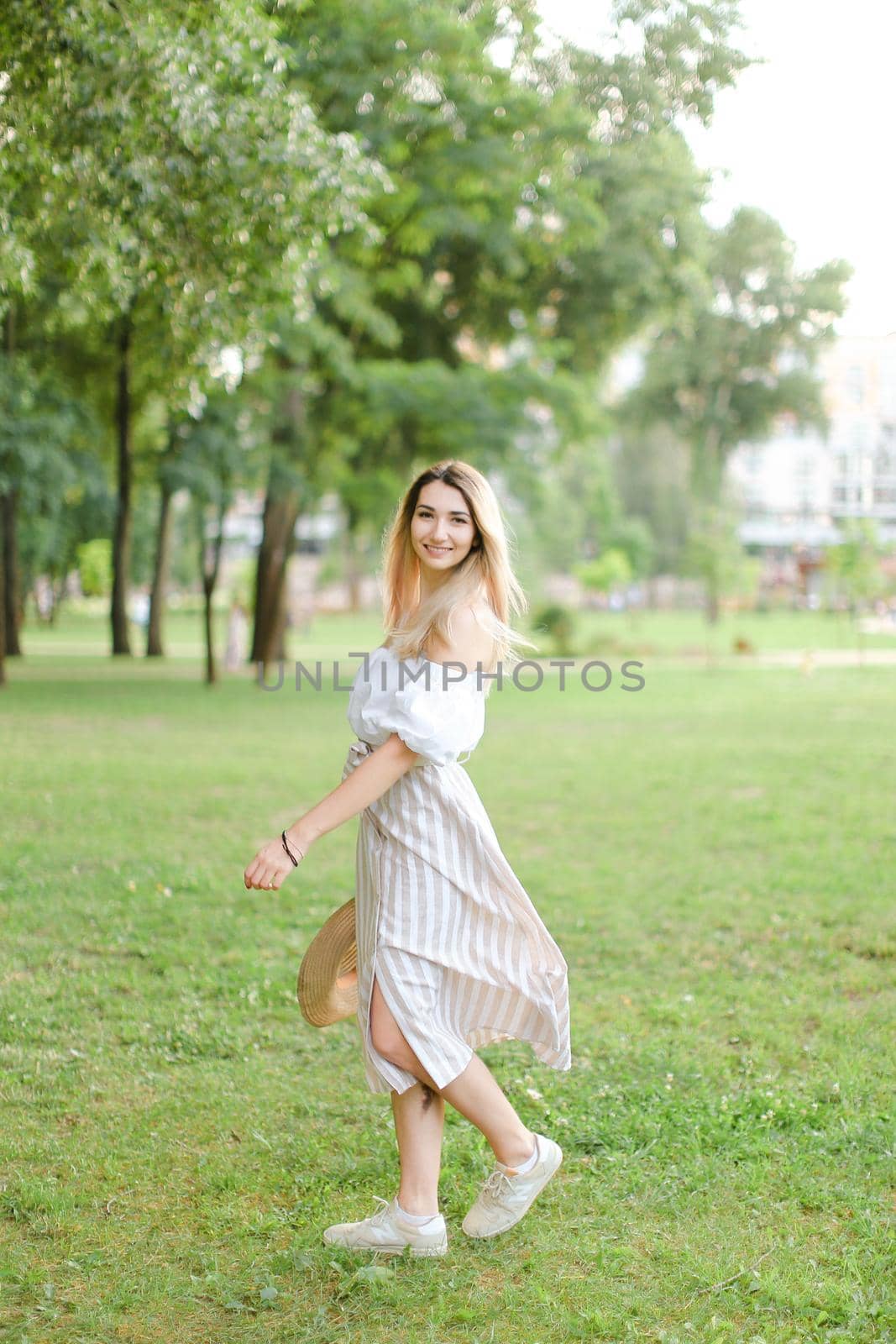 Young caucasian girl wearing dress, standing in park and keeping hat. Concept of summer season and fashion.