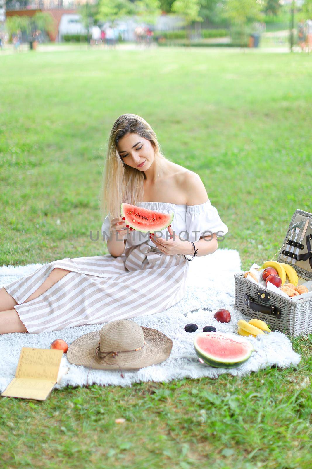 Girl sitting on plaid near fruits and hat, eating watermelon, grass in background. by sisterspro