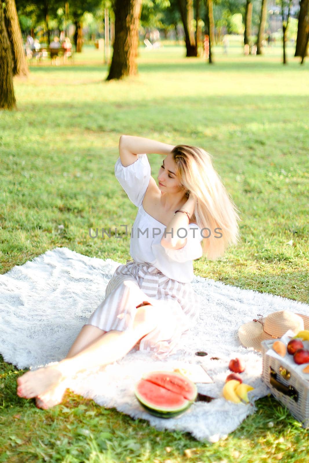 Young caucasian woman sitting on plaid with fruits in park. Concept of summer picnic, photo session in open air and vacations.