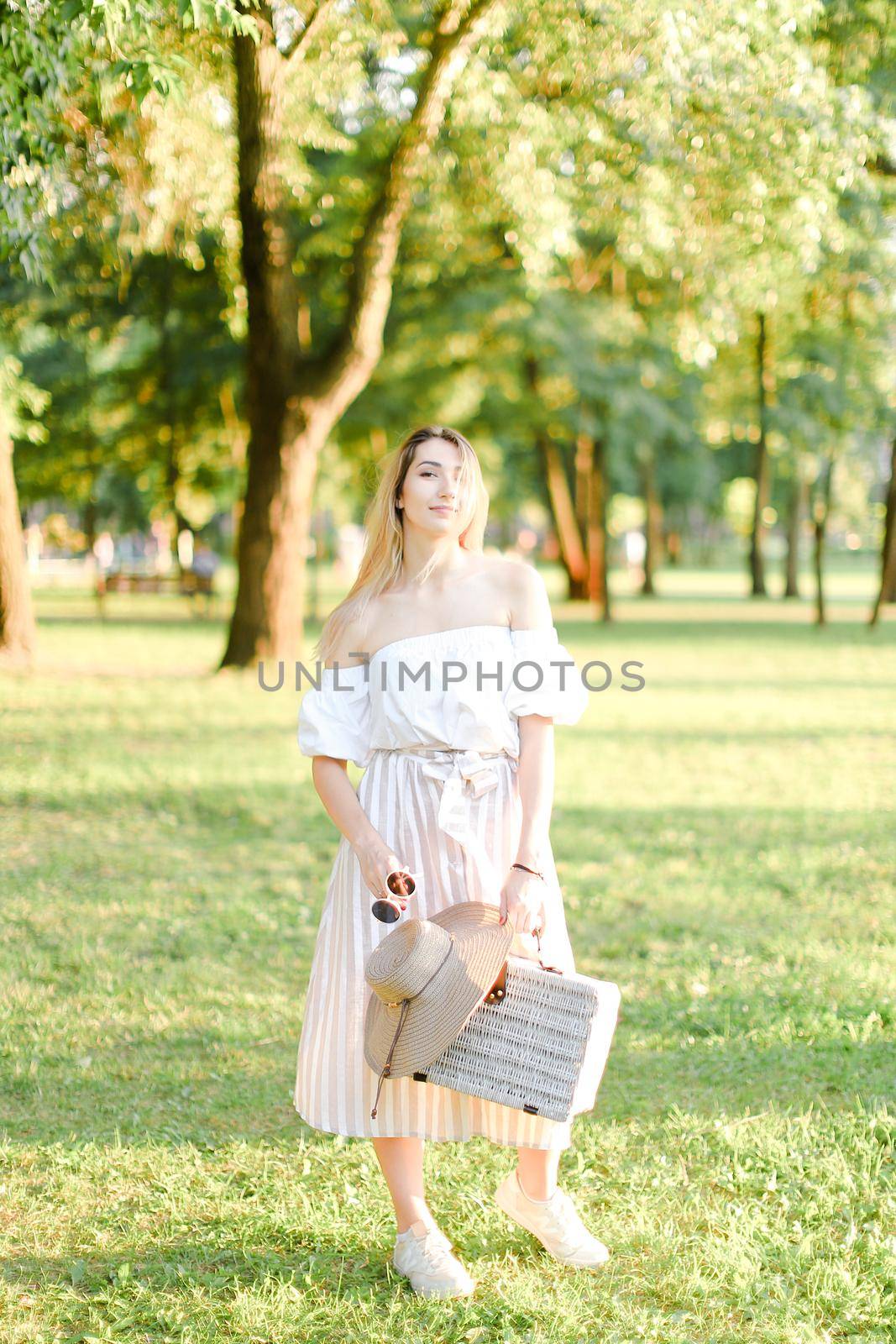 Young girl walking in garden and keeping bag, sunglasses and hat. Concept of walking in park and summer fashion.