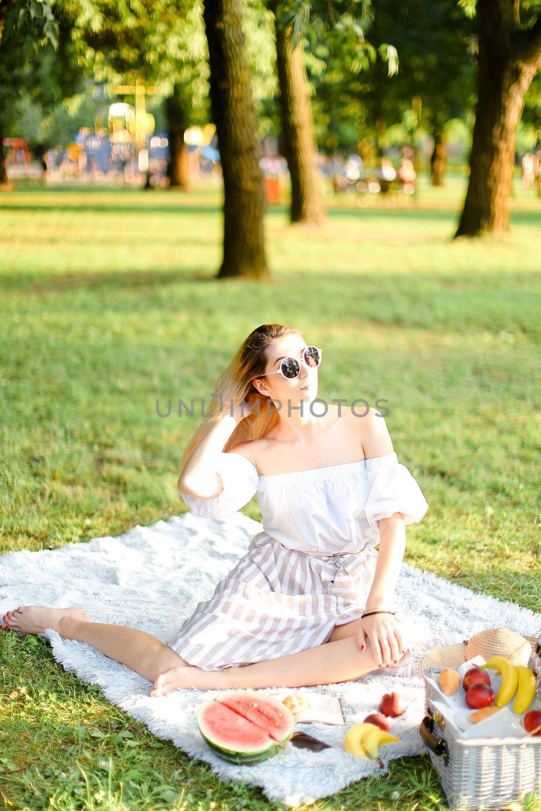 Young caucasian lady in sunglasses having picnic on plaid and sitting in park with fruits. Concept of resting in open air, leisure time and summer season