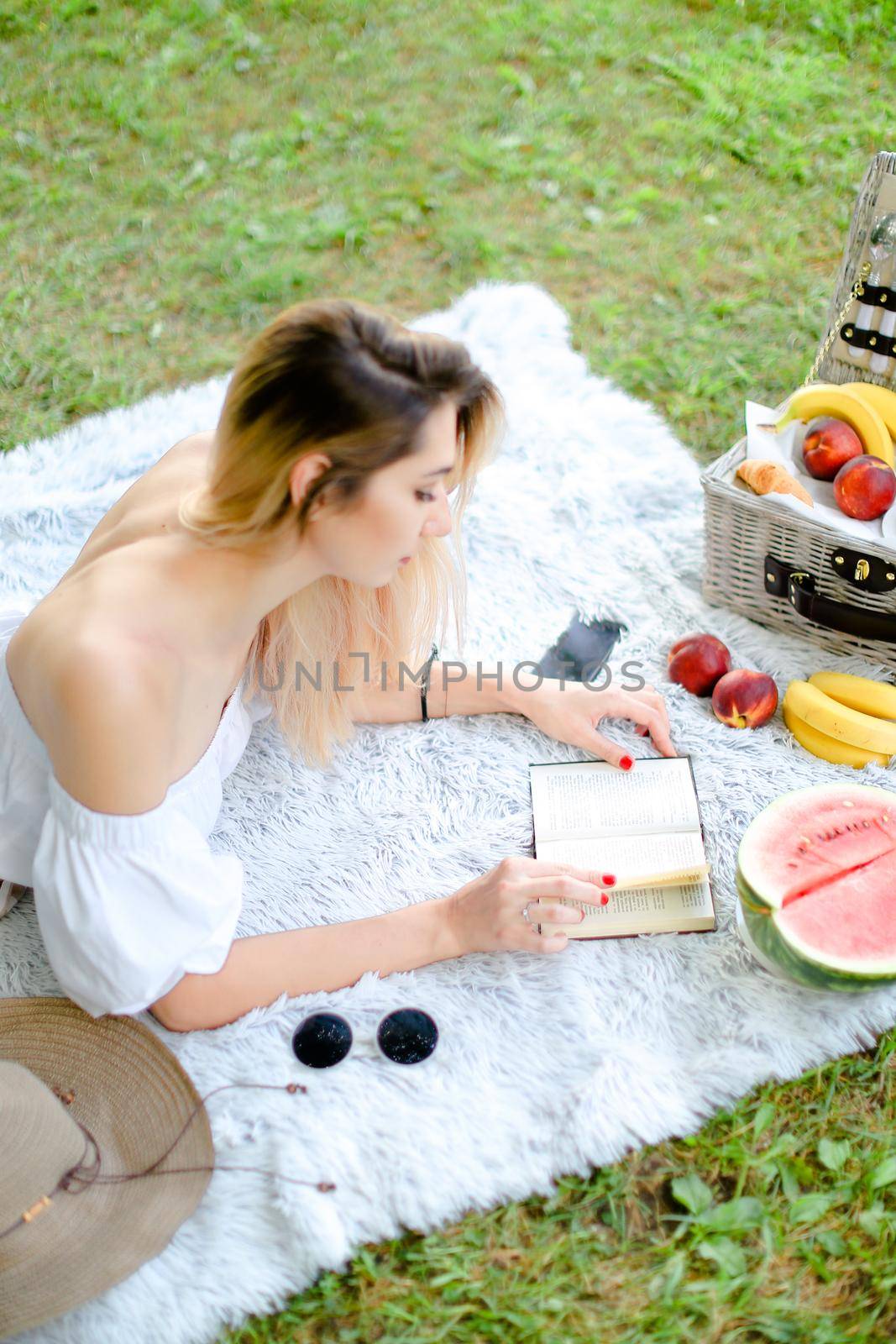 Young blonde woman reading book and lying on plaid near fruits and hat, grass in background. Concept of summer picnic and resting on weekends in open air.