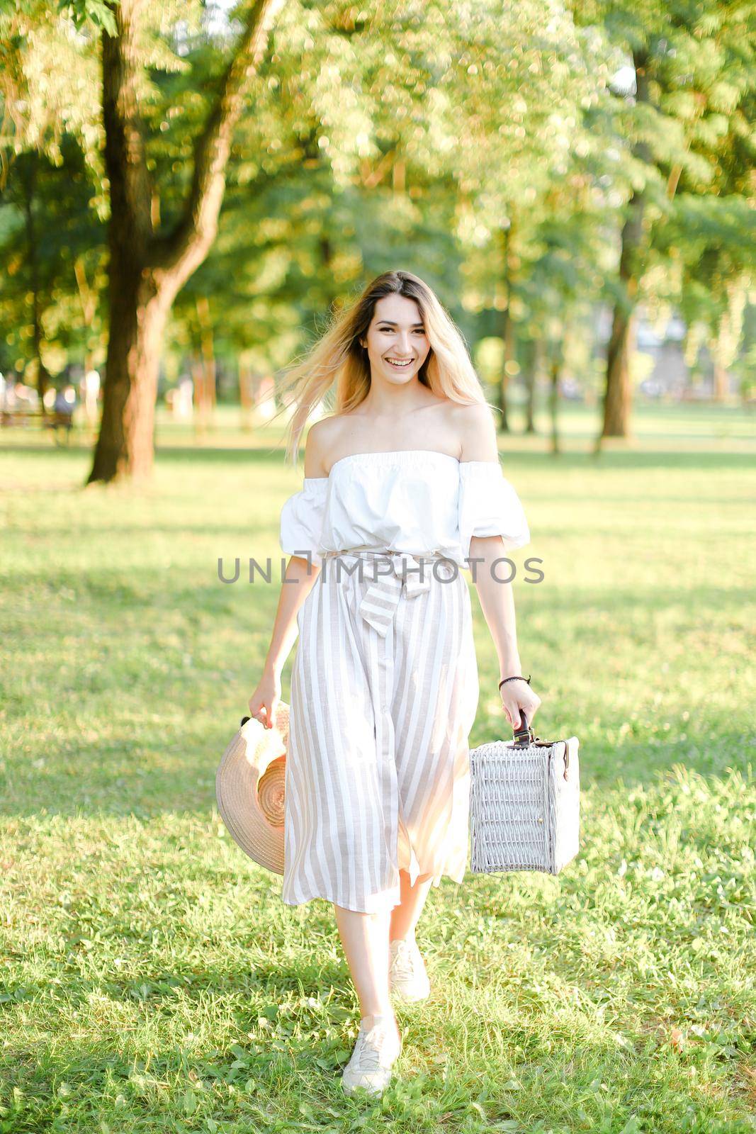 Young happy girl walking in garden and keeping bag, sunglasses and hat. Concept of walking in park and summer fashion.