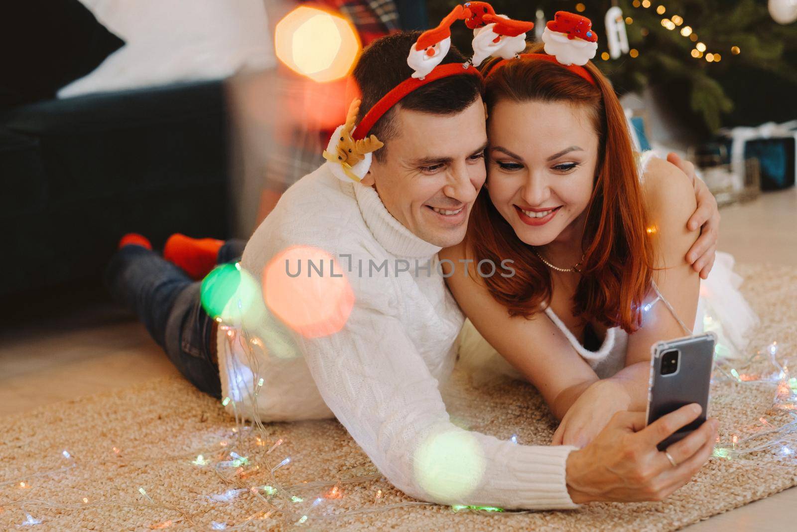 a happy married couple is lying on the floor at home near the Christmas tree and taking pictures of themselves.