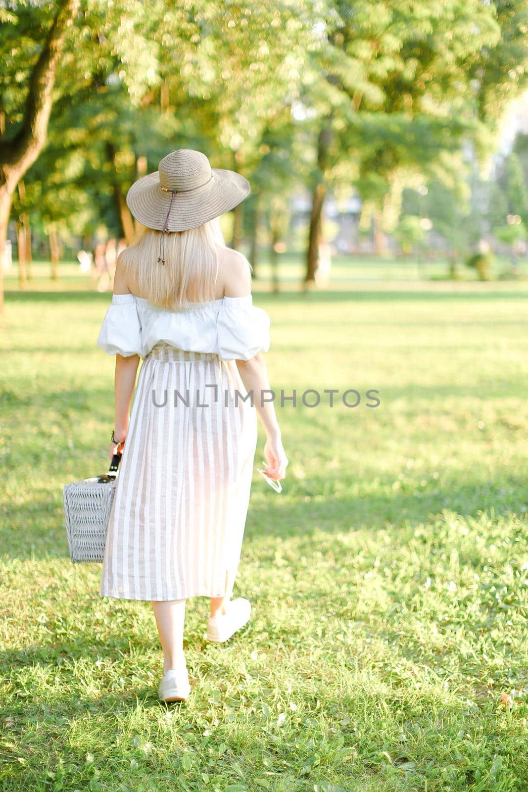 Back view of young pretty girl in hat walking in garden and keeping bag. Concept of walking in park and summer fashion garb.