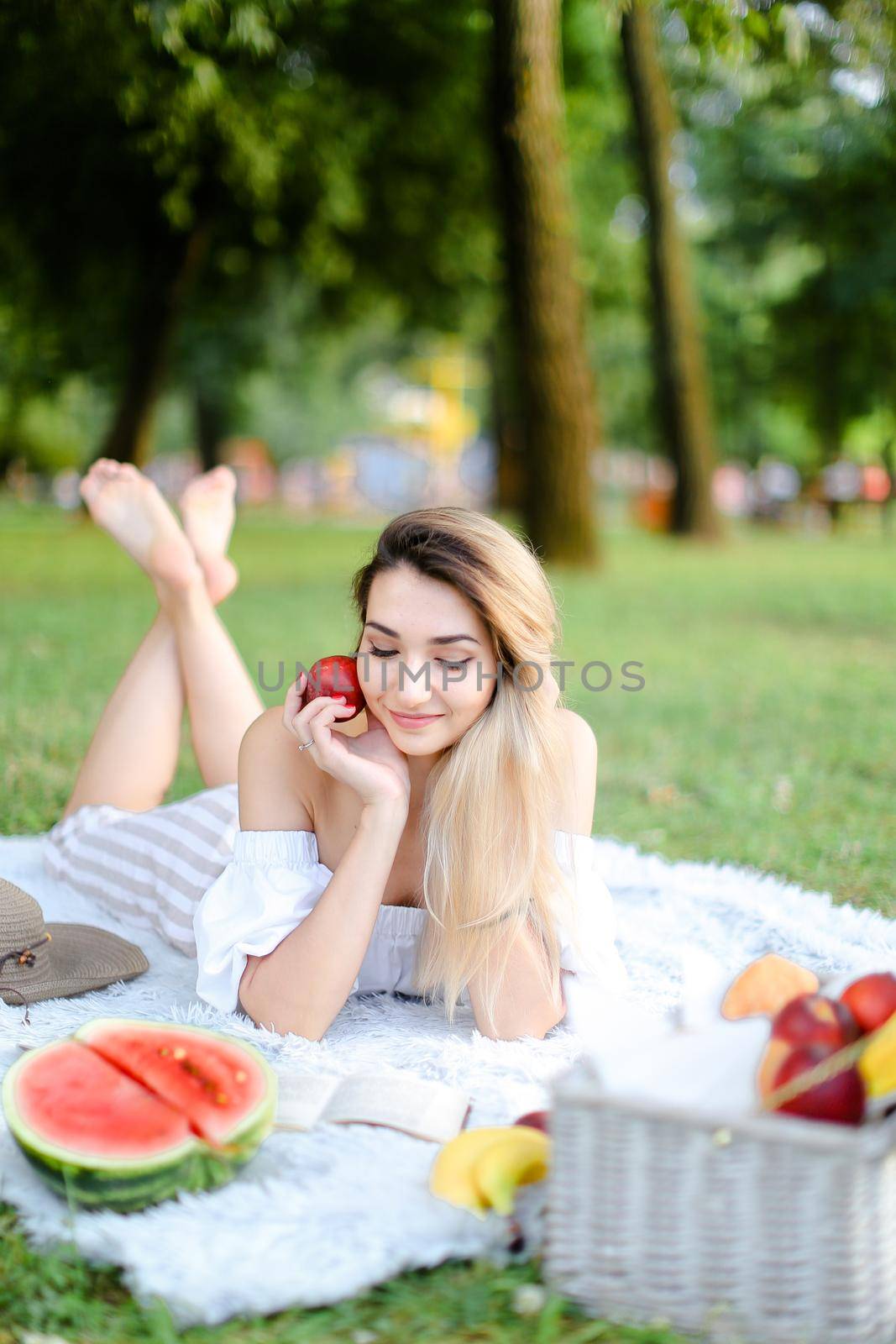 Young nice woman lying on plaid in park, reading book near watermelon and keeping apple. by sisterspro