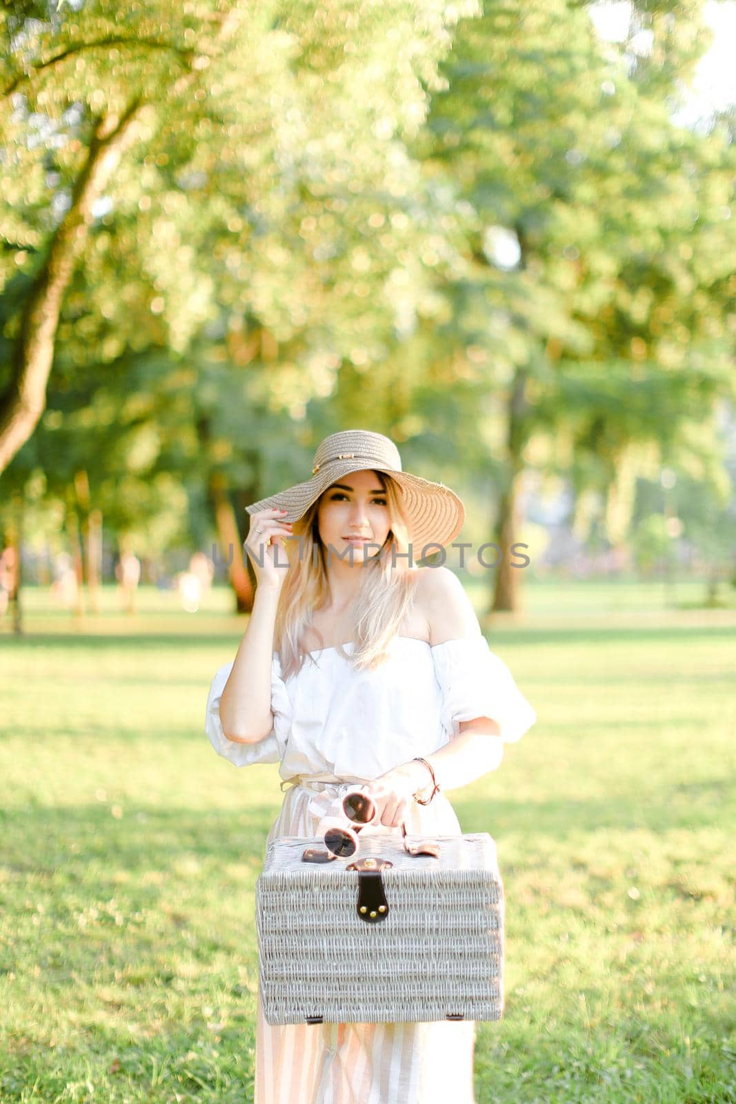 Young nice woman wearing hat and dress standing in garden with bag. Concept of beautiful female person, summer fashion and walking in park.