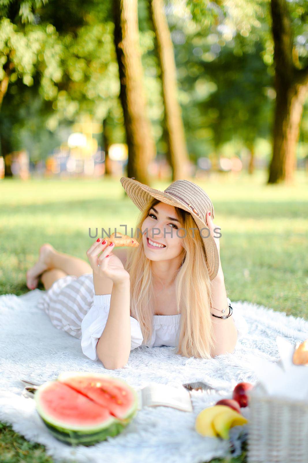 Young blonde girl in hat with croissant lying in park on plaid near watermelon. Concept of having free time, picnic and bakery products.