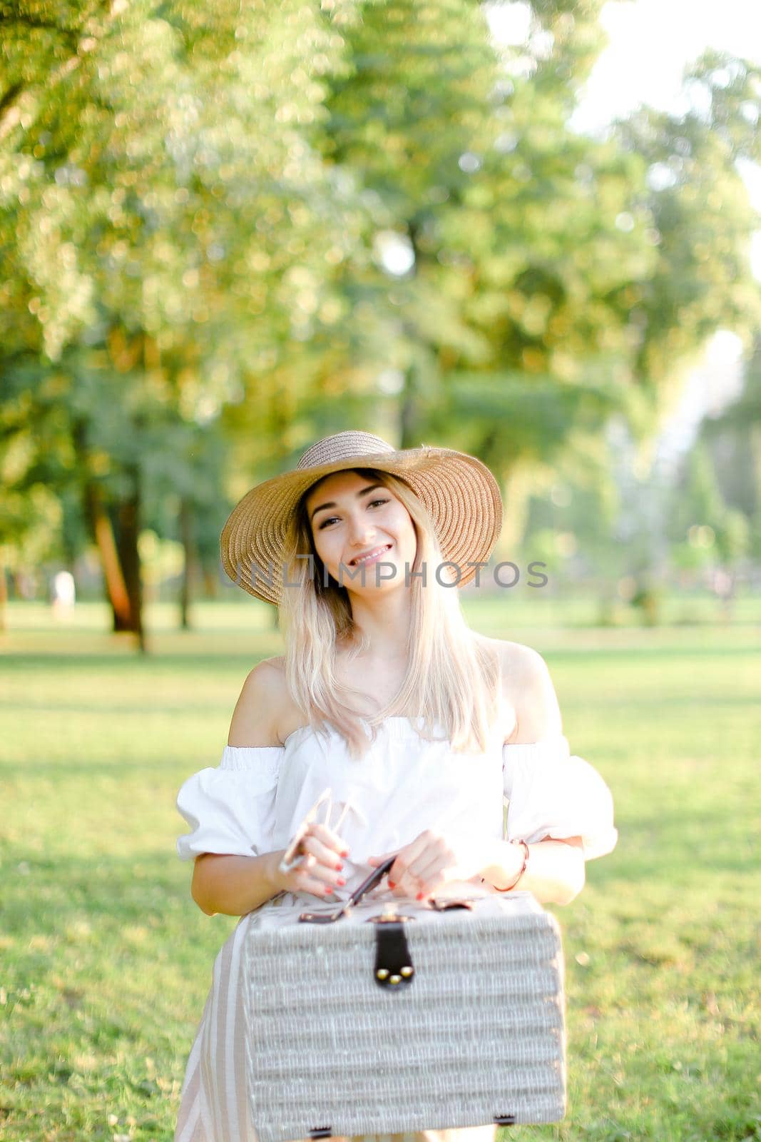 Young woman wearing hat and dress standing in garden with bag. Concept of beautiful female person, summer fashion and walking in park.