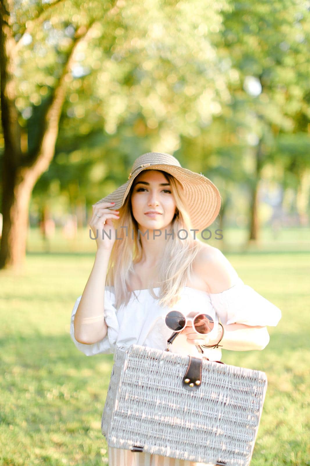 Young happy woman wearing hat and dress standing in garden with bag. Concept of beautiful female person, summer fashion and walking in park.