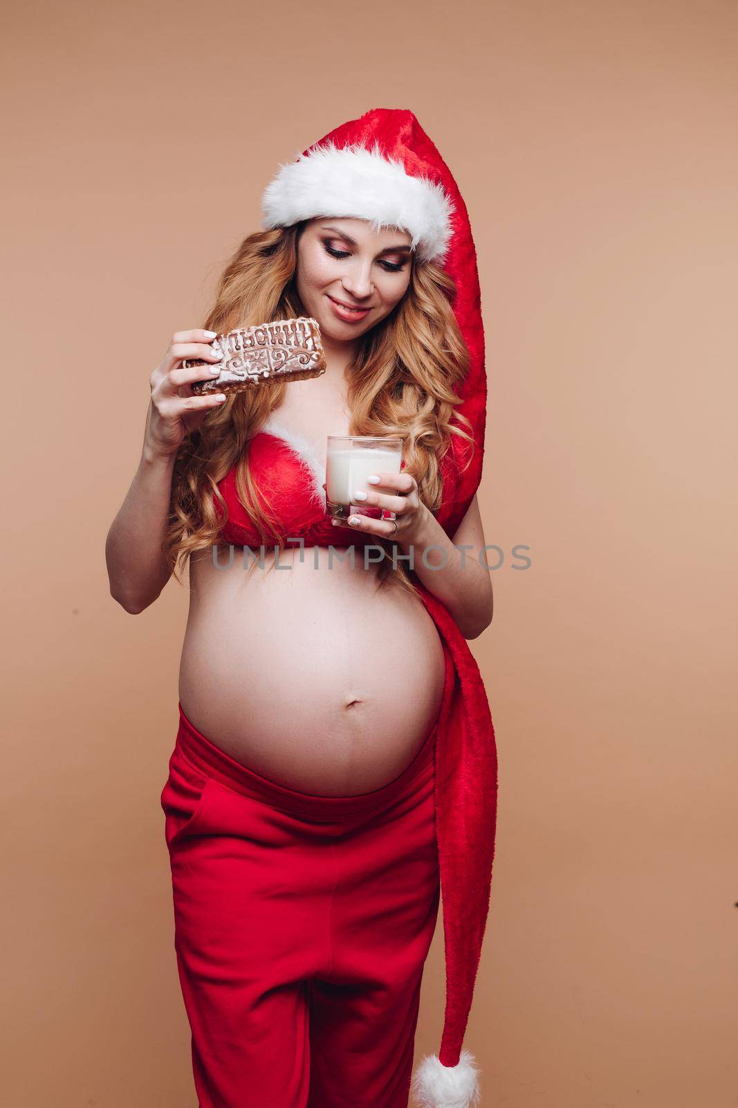 Attractive pregnant woman in a Santa costume eats gingerbread with milk in the studio