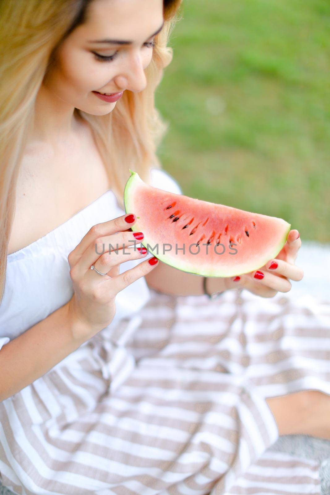 Young caucasian girl eating watermelon and wearing dress, grass in background. Concept of summer photo session, picnic.