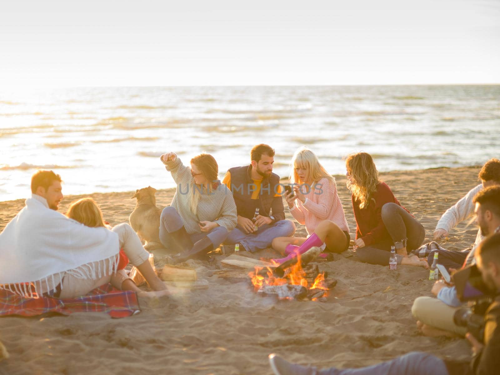 Happy Carefree Young Friends Having Fun And Drinking Beer By Bonefire On The Beach As The Sun Begins To Set