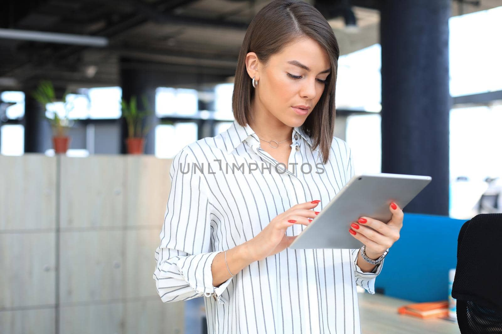 Portrait of young businesswoman looking at touch pad screen while standing in modern office space interior