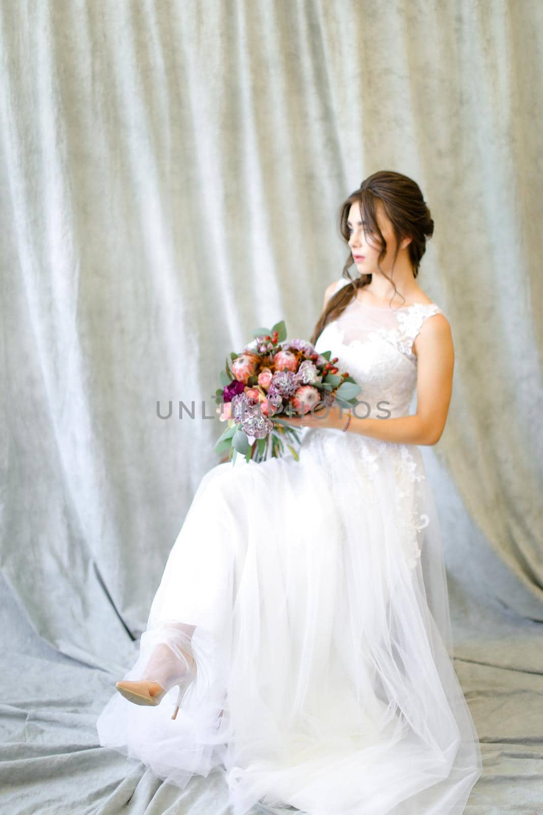 Caucasian brunette bride sitting at photo studio with bouquet of flowers. Concept of wedding and fianceee fashion.