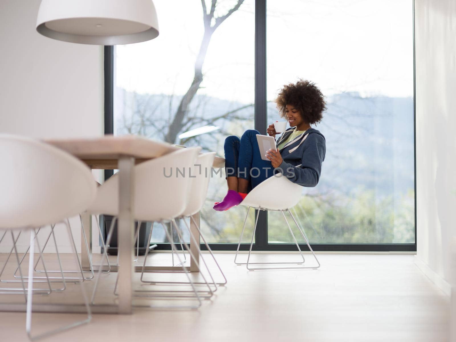 Young african american woman smiling sitting near bright window while looking tablet computer and holding white mug in her luxury home