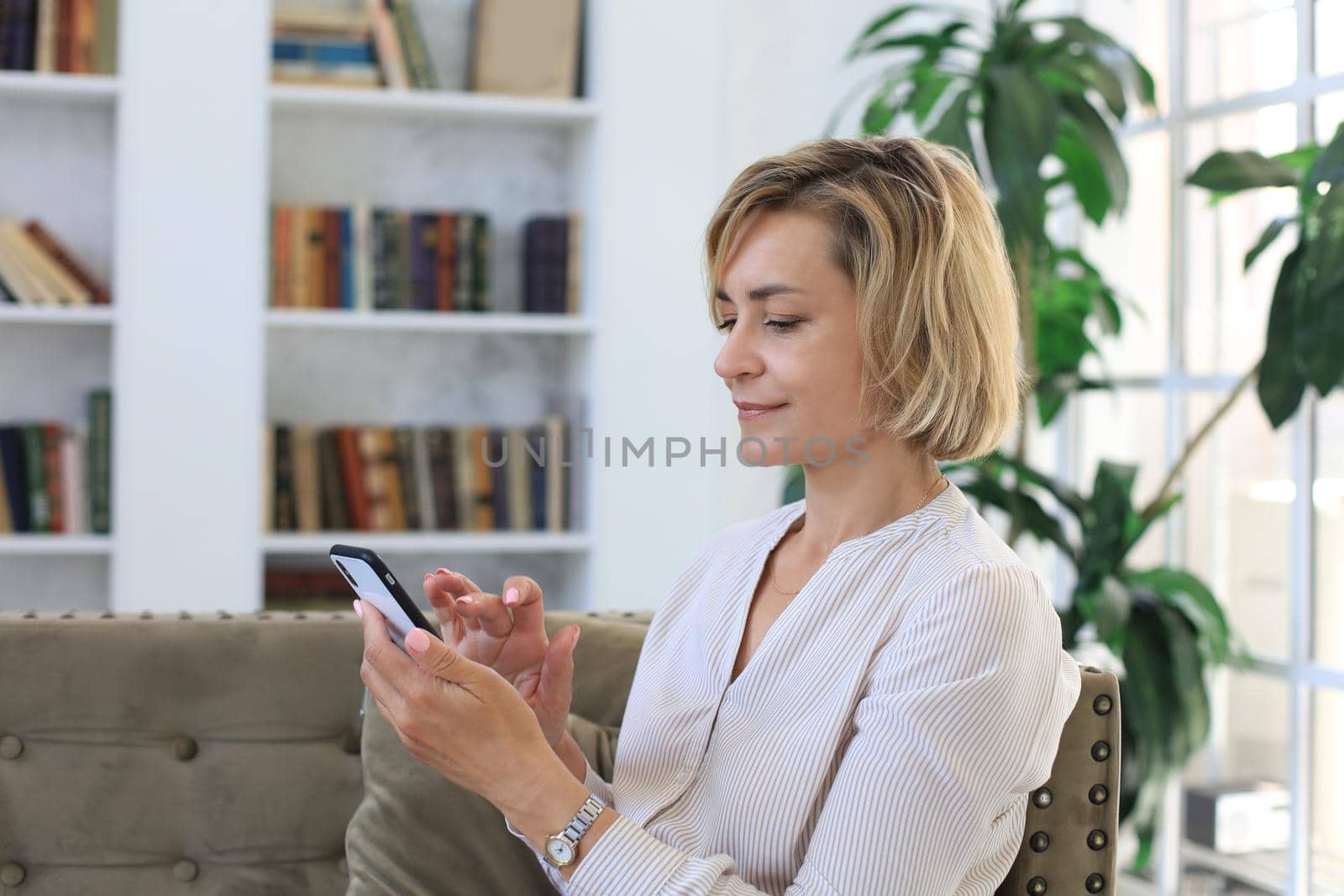 Smiling middle aged woman holding phone, using mobile device apps, looking at screen, while sitting on couch