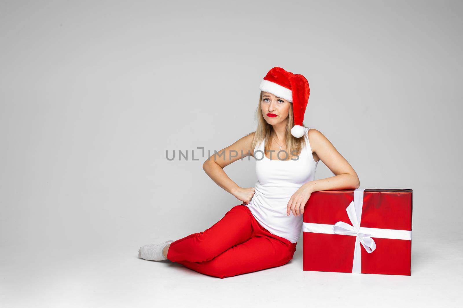 Stock photo of a sad Caucasian woman with blonde hair wearing red Santa hat sitting on the floor with Christmas present looking unhappy.