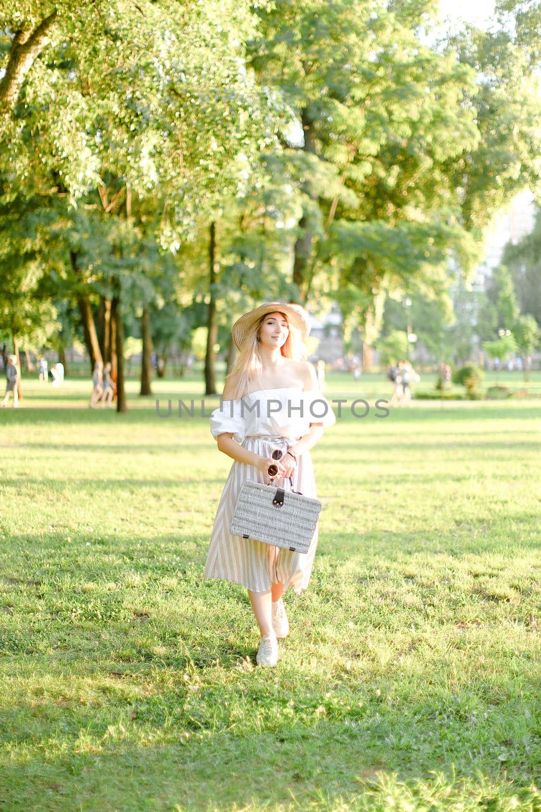 Young pretty girl walking in garden and keeping bag, sunglasses and hat. Concept of walking in park and summer fashion garb.