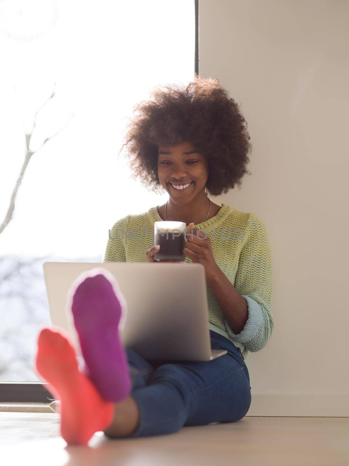 Young african american woman smiling sitting on the floor near bright window while looking at open laptop computer and holding mug at home