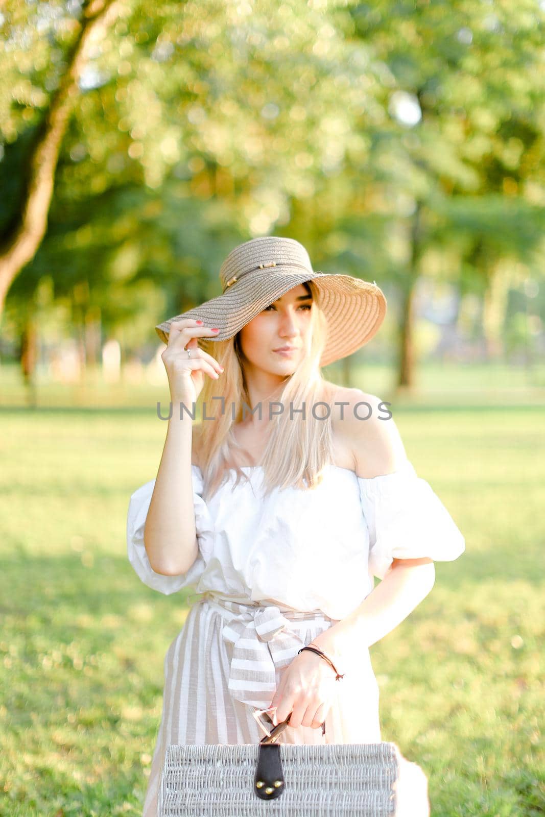 Young caucasian woman wearing hat and dress standing in garden with bag. Concept of beautiful female person, summer fashion and walking in park.