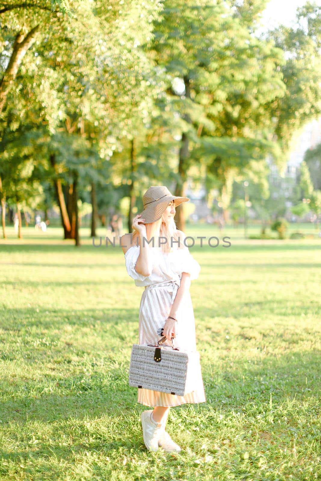 Young blonde american woman wearing hat and dress standing in garden with bag. Concept of beautiful female person, summer fashion and walking in park.