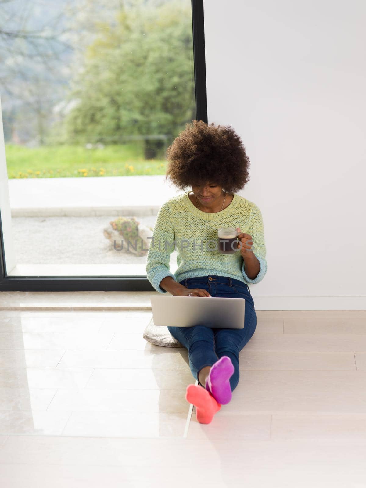 Young african american woman smiling sitting on the floor near bright window while looking at open laptop computer and holding mug at home