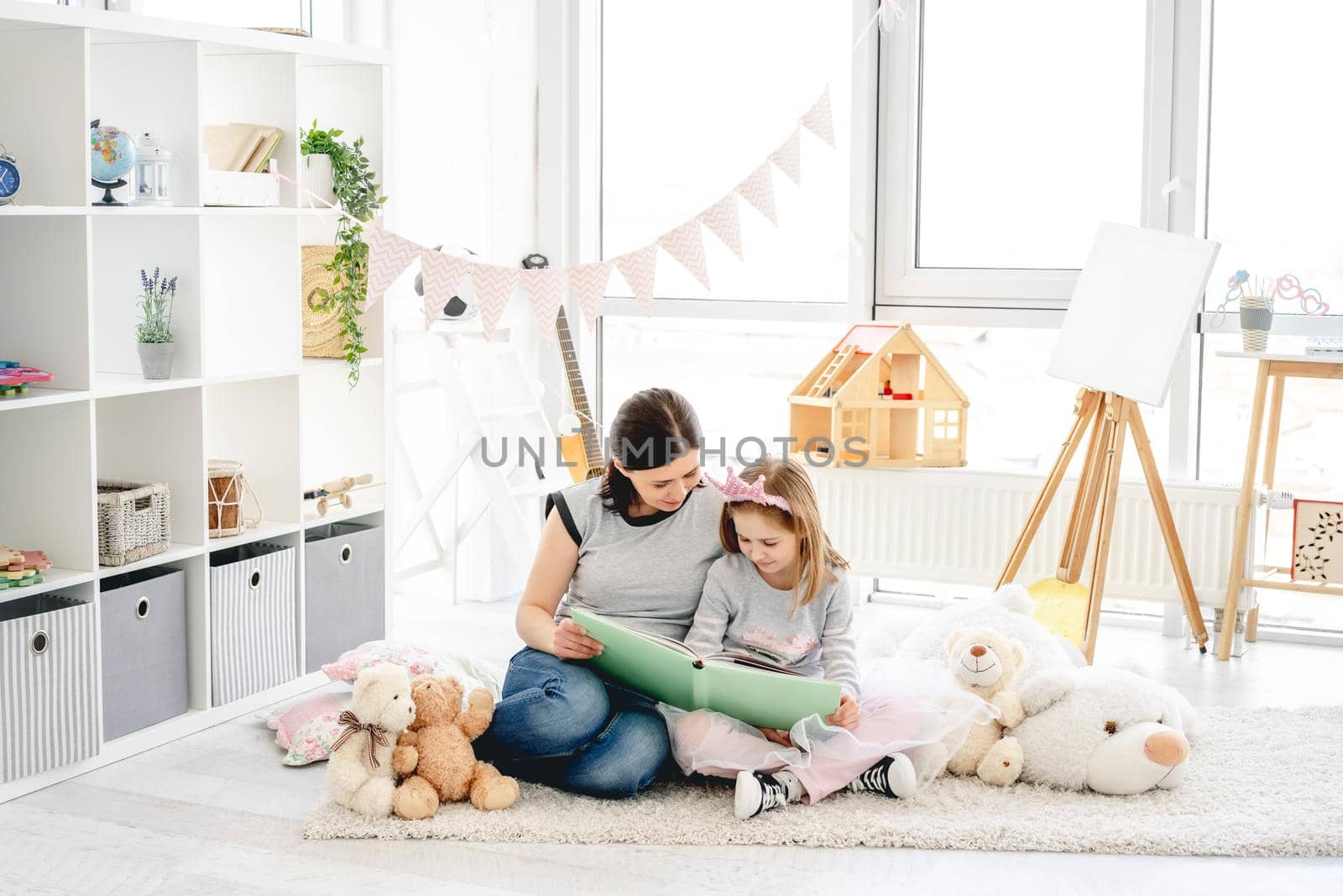 Beautiful mother and daughter reading book sitting on floor in kids room