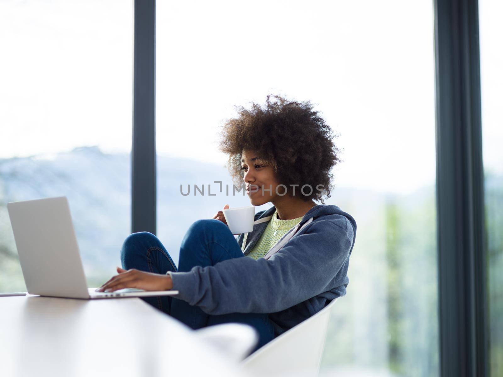 Young african american woman smiling sitting near bright window while looking at open laptop computer on table and holding white mug in her luxury home