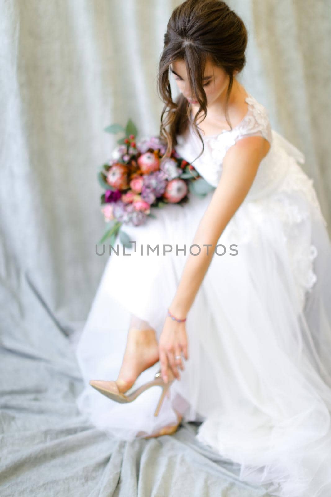 Young brunette bride with flowers putting on shoes at photo studio. by sisterspro