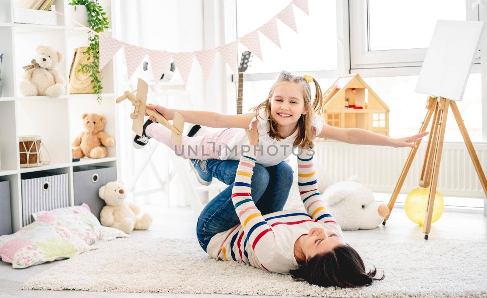 Laughing daughter flying on mother's legs in bright room