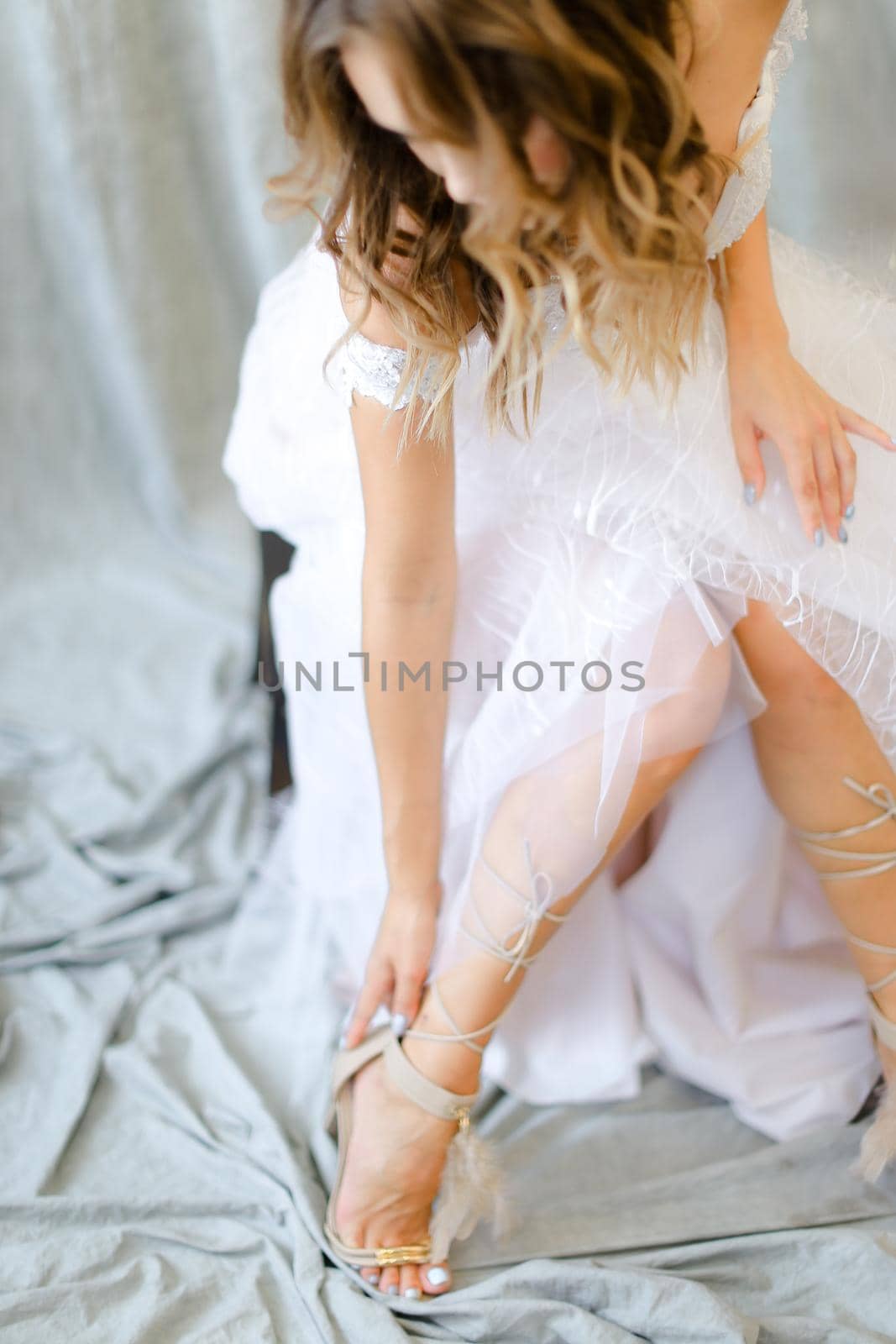 Young blonde caucasian bride in white dress putting on shoes at photo studio. by sisterspro