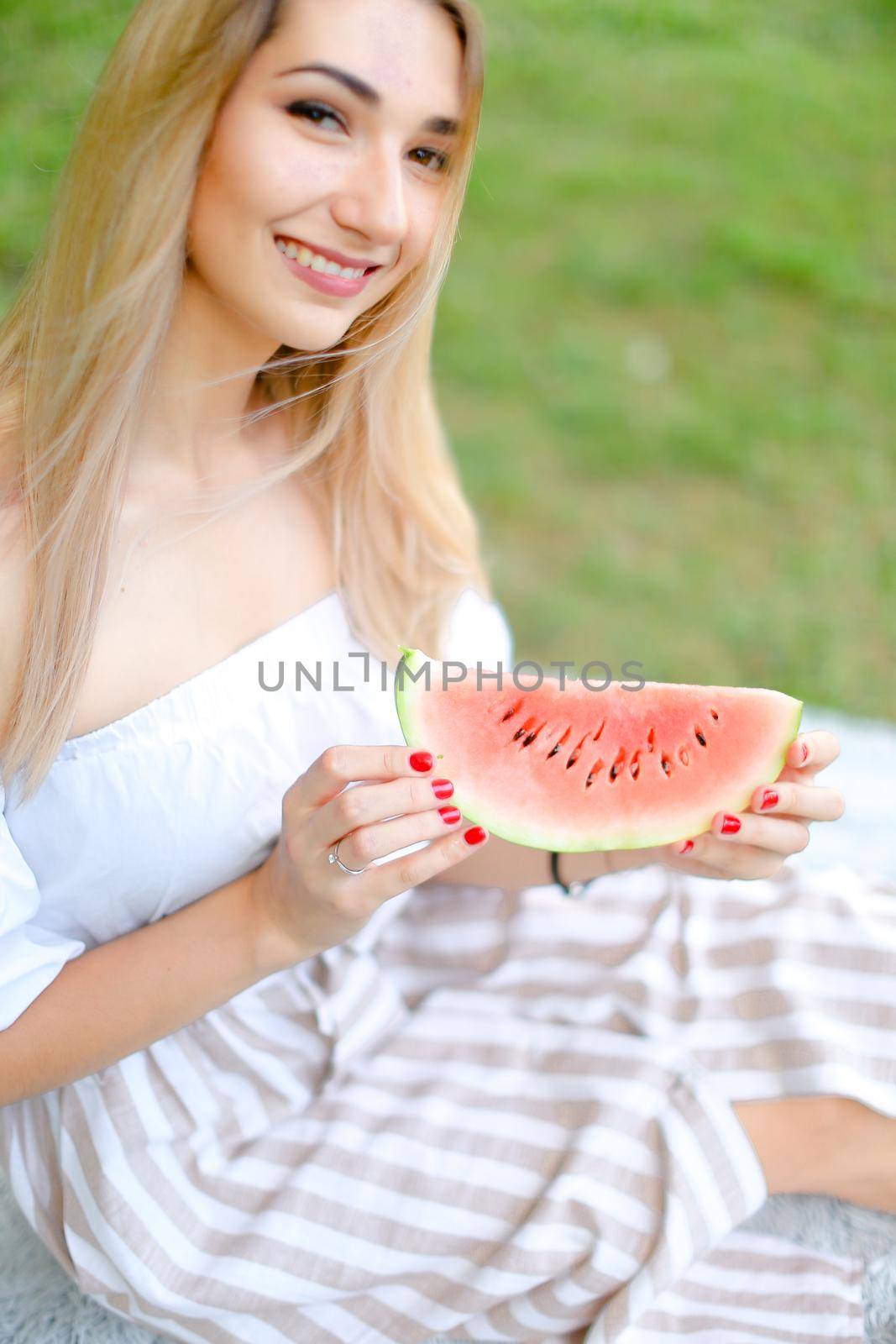 Young american girl eating watermelon and wearing dress, grass in background. Concept of summer photo session, picnic.