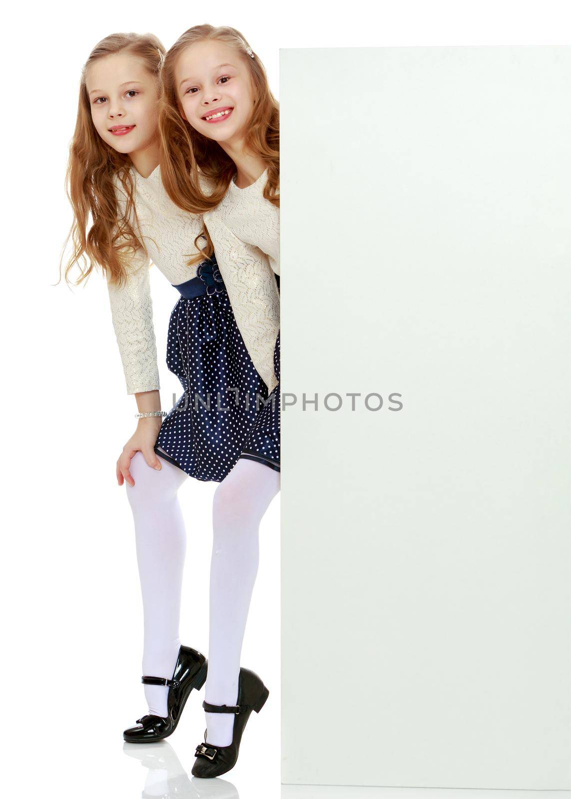 Two cute little girls look out from behind a white advertising banner. On the banner you can make any advertising inscription or put a picture or logo. It is isolated on a white background.