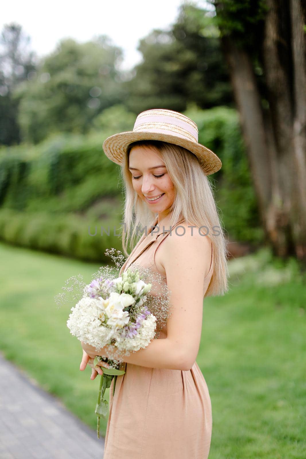 Young girl standing in yeard with bouquet of flowers and wearing hat with overalls. Concept of yuoth and summer season, fashion.