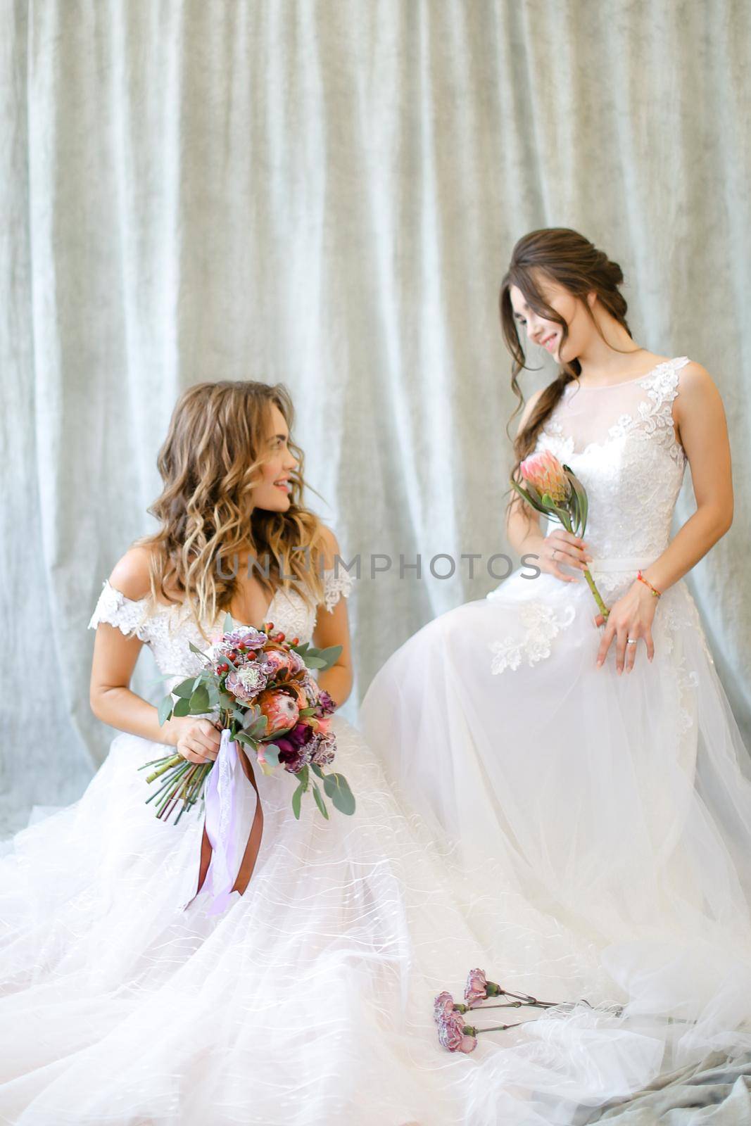 Two brides wearing white dress and sitting at studio with flowers. Concepr of bridal photo session and wedding.